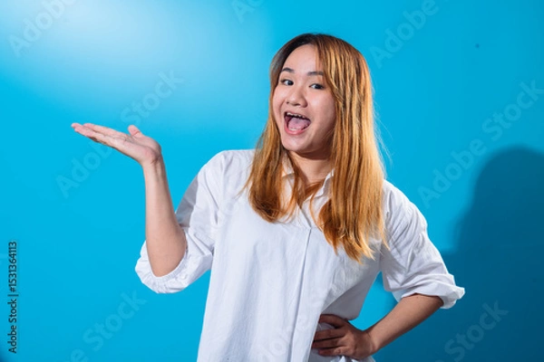 Fototapeta Asian woman with long straight hair in white shirt smiling while holding one hand open to the side as if presenting or showing something, standing confidently against a solid blue studio background