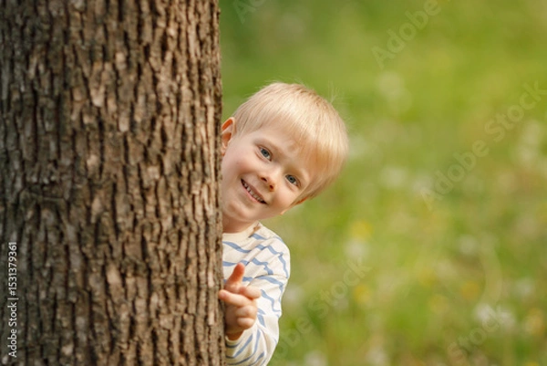 Obraz A cheerful blonde-haired boy with bright blue eyes playfully peeks out from behind a textured tree trunk, offering a sweet, engaging smile directly at the viewer.