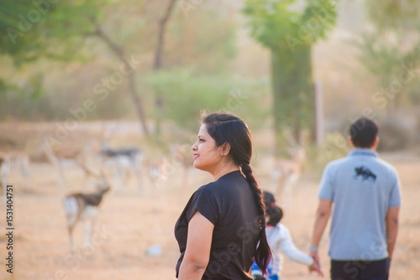 Obraz Beautiful Indian lady in black t-shrit with blurred background showing a man holding a child's hand and a herd of blackbucks in the grassland Tal Chhapar Wildlife Sanctuary. 
