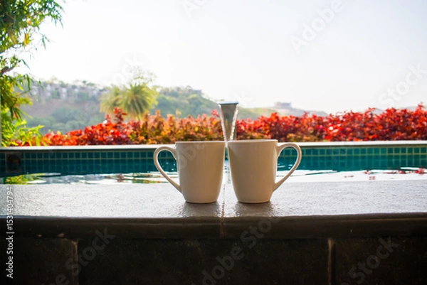 Obraz Two coffee mugs placed together with a blurred background of a serene swimming pool, symbolizing a romantic getaway or honeymoon vacation in India. couple holidays, tropical retreats, or leisure
