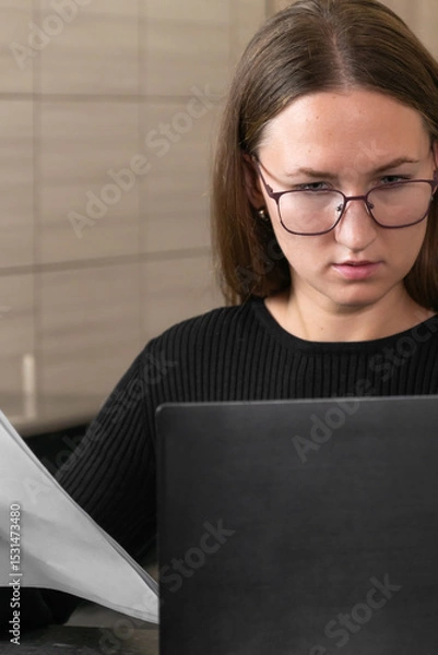 Obraz Young woman accountant wearing eyeglasses working from home, reviewing financial documents and using laptop, performing accounting tasks remotely. Vertical photo