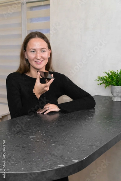 Obraz Smiling young woman recording a voice message on her smartphone while comfortably leaning against the kitchen counter in her stylish modern apartment, enjoying a casual moment of connection.