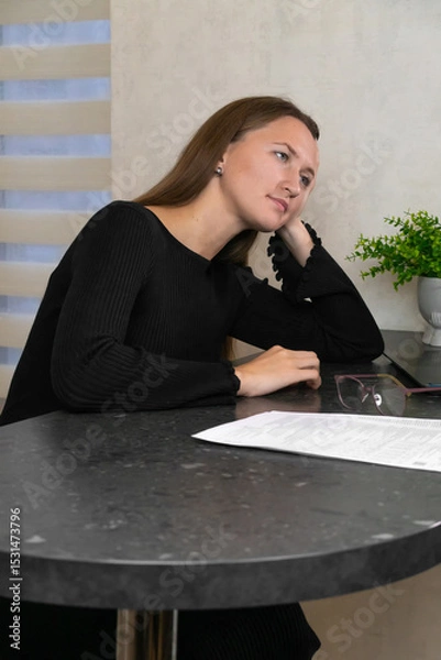 Obraz Young businesswoman in black clothes sitting at table with documents, glasses and laptop, leaning on her hand and looking away, feeling tired and bored. Vertical photo