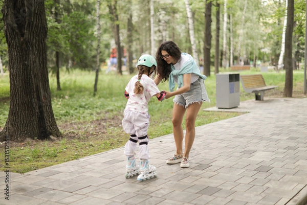 Fototapeta Mother teaching her daughter how to rollerblade in a park. The girl wears protective gear and helmet. A joyful and bonding outdoor moment filled with love and support.