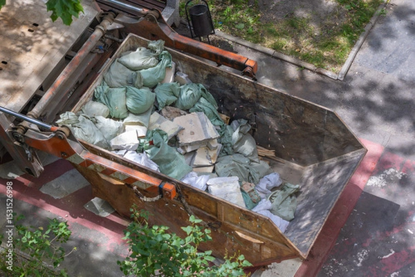 Fototapeta Top view of a construction dumpster filled with green bags containing building debris and rubble, surrounded by trees casting shadows on a sunny day