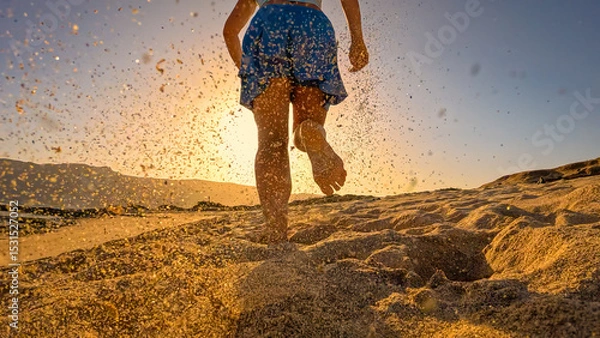 Fototapeta LOW ANGLE VIEW, LENS FLARE: Sporty woman in skort on an evening run along a sandy ocean shore. At sunset, she runs barefoot on beach in blue sportswear, leaving flying sand behind in golden light.