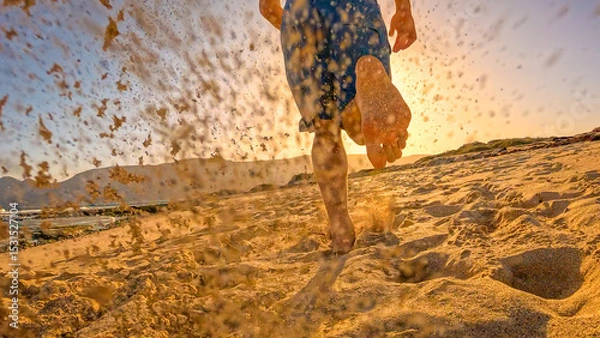 Fototapeta LOW ANGLE VIEW, LENS FLARE: Athletic man in blue shorts runs barefoot along sandy ocean beach in golden light. He is on an early morning run and enjoys relaxing and active holidays at Lanzarote island