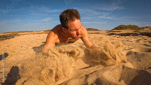 Fototapeta LOW ANGLE VIEW: Shirtless man falls forward onto the sand while running to the beach. Hands slam down and scatter sand into the air as late afternoon sunlight creates a dramatic, energetic atmosphere.