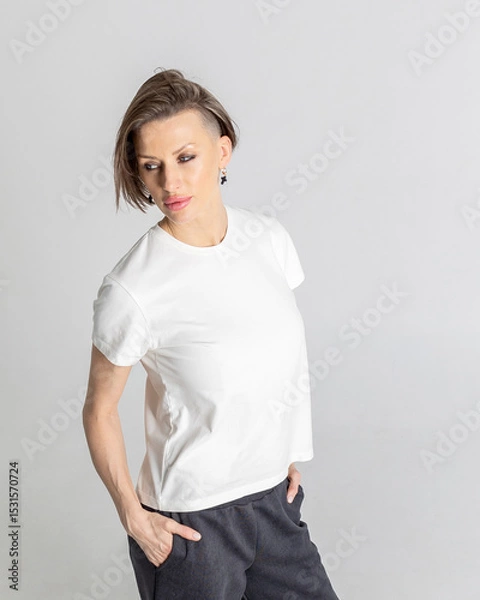 Obraz Portrait beautiful woman of model appearance. Girl in white T-shirt, black trousers on grey background. Pretty female with trendy makeup posing in studio. Vertical photo.