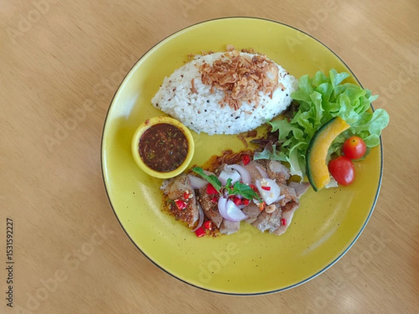Fototapeta top view, Waterfall pork, fried garlic on rice and nam jim jaew in a yellow bowl on a yellow plate on a wooden background.