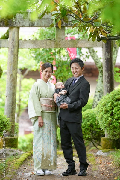Fototapeta On a spring day in May before a green torii gate, a one-month-old Japanese baby in hakama is held by a suited man, walking with a kimono-clad mother in their twenties, during a gentle family stroll.