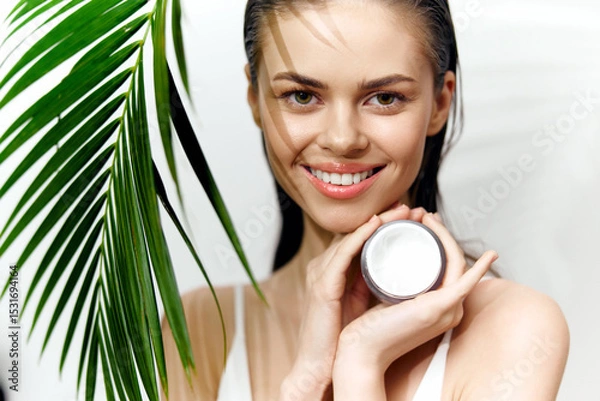 Fototapeta Smiling model holding skincare cream with tropical greenery background, showcasing beauty and freshness in wellness concept.
