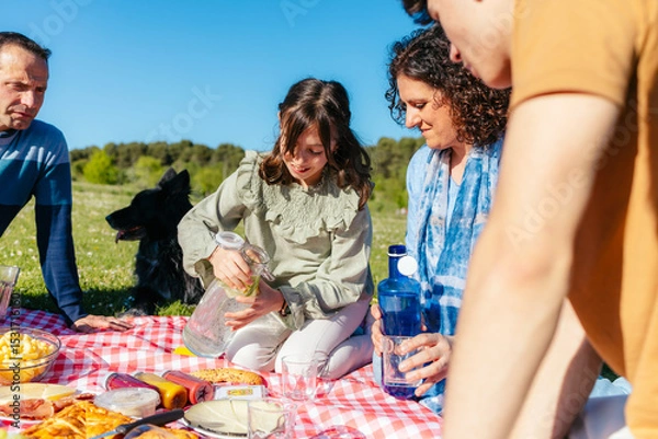 Fototapeta Happy family having fun, eating and playing with their dog during a picnic in a park, girl is pouring lemonade