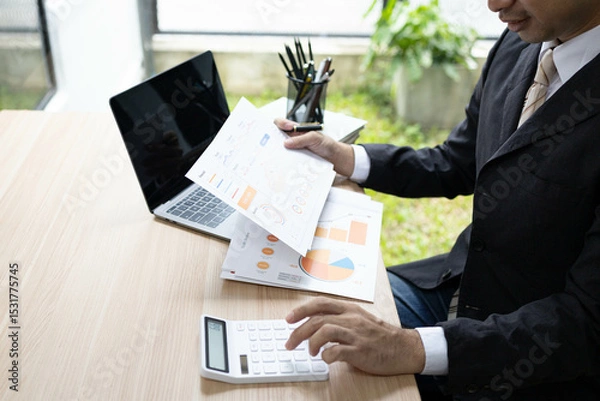 Fototapeta A man in a suit is sitting at a desk with a laptop and a calculator. He is holding a piece of paper and he is working on some financial documents. Concept of professionalism and focus
