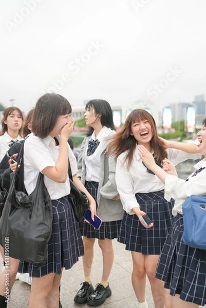 Fototapeta In early summer in Shanghai, near The Bund, Japanese high school girls in uniforms chat and laugh in a busy tourist area, their cheerful presence adding youthful charm to the city's historic vibe.