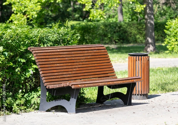 Fototapeta A wooden bench sits in a park next to a trash can