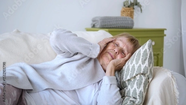 Fototapeta Elderly woman experiencing a severe headache, lying on a comfortable sofa with her hands on her temples, seeking relief from the pain in a cozy home environment