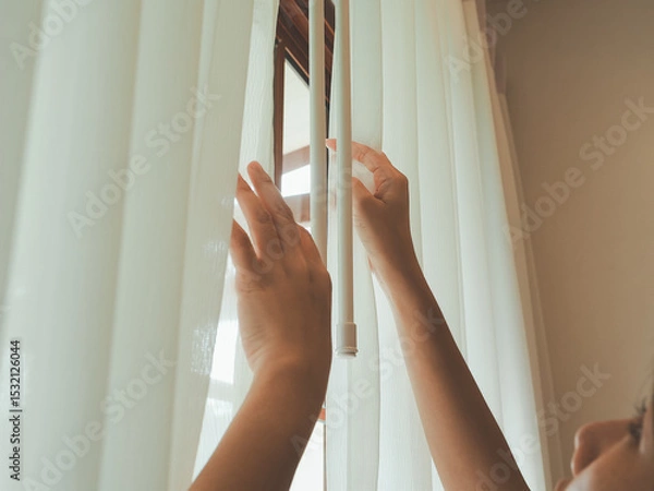 Fototapeta A person gently adjusts white vertical blinds on a window, creating a soft and calming atmosphere with natural light filtering through the fabric.