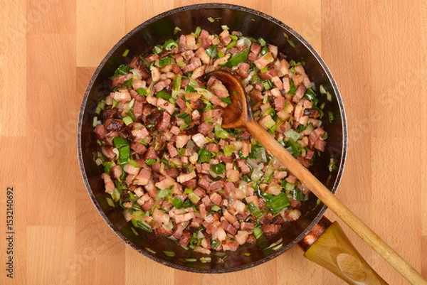 Obraz A top-down view shows diced pancetta and chopped green onions being stirred in a dark skillet with a wooden spoon