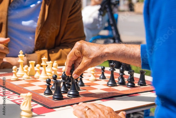 Obraz A casual chess game between two individuals, one in a blue sweater and the other in a brown jacket, with sunlight and outdoor seating in the background.