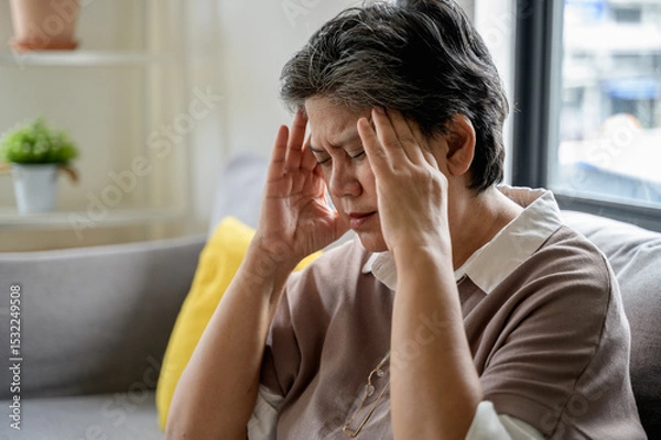 Fototapeta An elderly Asian woman is sitting on a sofa at home, experiencing a headache, dizziness, and a migraine. She looks unwell and uncomfortable. Senior health care daily lifestyle problem concept.