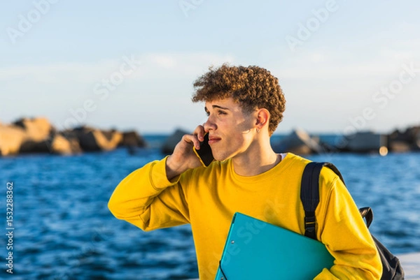 Fototapeta College student talking on smartphone by the sea holding a binder