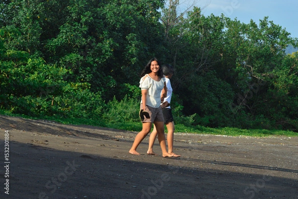 Fototapeta With sandals in hand and smiles on their faces, the girls enjoy a slow stroll on black sand surrounded by tropical greenery and morning sunshine
