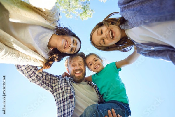 Fototapeta Bottom view of happy young family of four looking cheerful at camera outdoors on blue sky background. Portrait of smiling mother, father and children enjoying time in nature together.