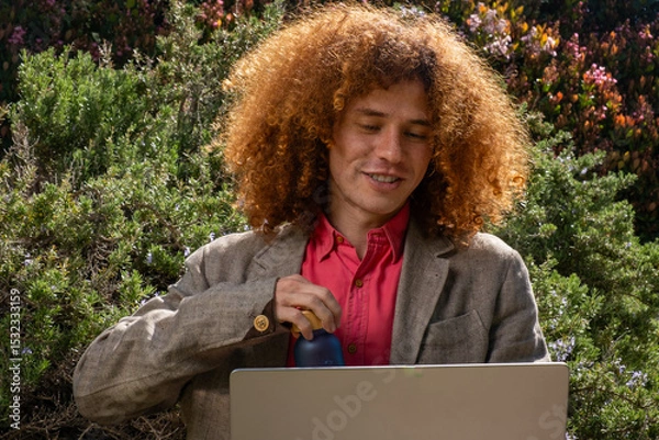 Fototapeta Young man with curly hair working on a laptop outdoors, holding a reusable bottle. Concept of sustainable remote work, eco-friendly lifestyle and digital wellbeing.