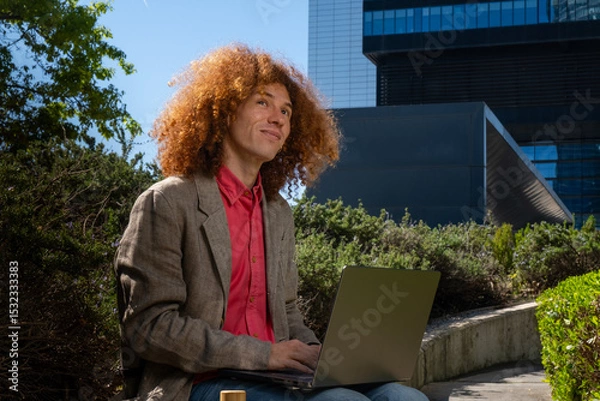 Fototapeta Young man with curly hair working on a laptop outdoors, looking up with a thoughtful expression. Concept of sustainable remote work, creativity, eco-friendly business and future vision.