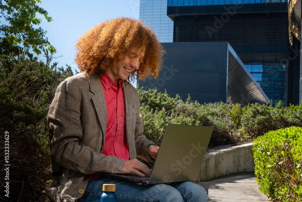 Fototapeta Young man with curly hair smiling while working on a laptop outdoors, reusable bottle in foreground. Concept of happy remote work, eco-friendly business and sustainable digital lifestyle.