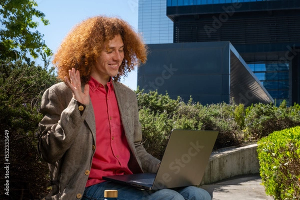 Fototapeta Young man with curly hair waving during a video call outdoors, working on a laptop with reusable bottle visible. Concept of remote communication, sustainable digital work and eco-friendly lifestyle.