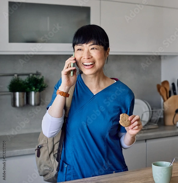 Fototapeta Portrait of a young asian nurse woman gettyng ready for work drinking a glass of water for hydration and talking on a mobile phone smartphone  standing in kitchen home. Overworked, busy, stress, urgen