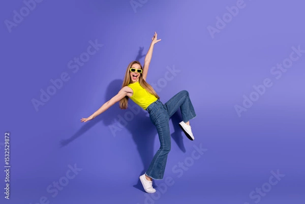 Fototapeta Joyful young woman dancing with enthusiasm against a bright purple background wearing a stylish yellow top and sunglasses
