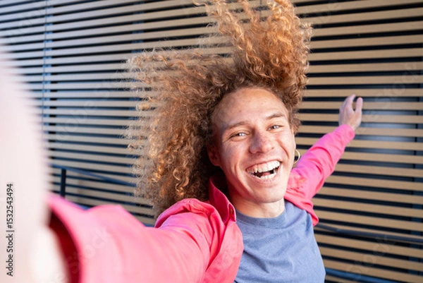 Fototapeta Cheerful young man with curly hair taking dynamic selfie outdoors in pink jacket. Joyful student, social media lifestyle, self expression and youthful energy concept.