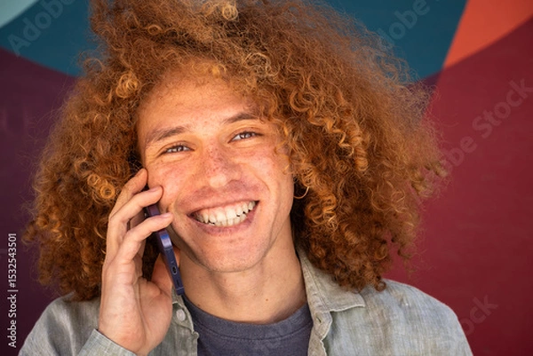 Fototapeta Close-up portrait of smiling young man with curly hair talking on smartphone against colorful wall. Cheerful student, modern communication, youthful lifestyle and personal branding concept.