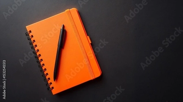 Fototapeta Orange Notebook and Pen: A vibrant orange notebook with a black pen rests on a dark gray surface, creating a minimalist yet striking image perfect for representing organization, planning.