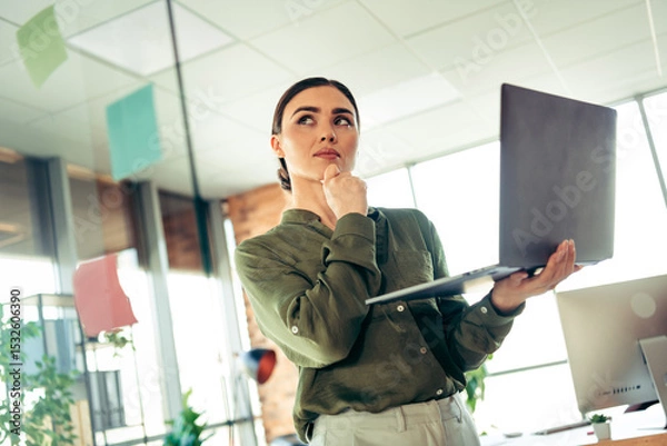 Fototapeta Thoughtful businesswoman holding laptop in a professional modern office space considering ideas for a corporate project
