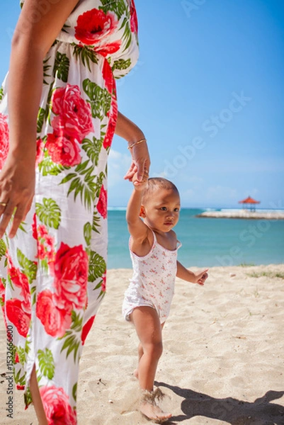 Fototapeta Asian mother in floral dress holding the hand of her toddler girl, helping her walking barefeet on beach sand under harsh sunlight on hot sunny summer day with blue ocean and blue sky on background