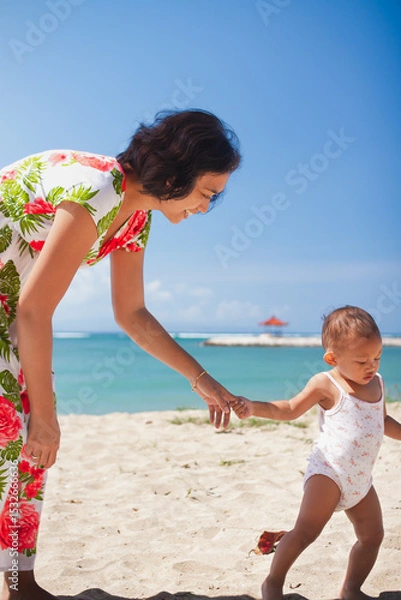 Fototapeta Asian mother in floral dress holding the hand of her toddler girl, helping her walking barefeet on beach sand under harsh sunlight on hot sunny summer day with blue ocean and blue sky on background