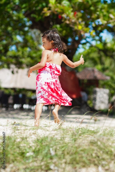 Fototapeta Cheerful Asian little girl in colorful casual tropical dress running and jumping around on the beach with a big smile on her face