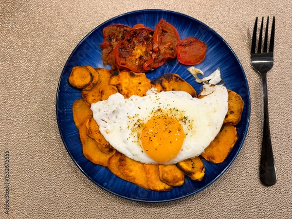 Fototapeta Breakfast meal consisting of fried sunny side up egg, sliced tomato, and yam sweet potato on blue dish setting in flat lay view