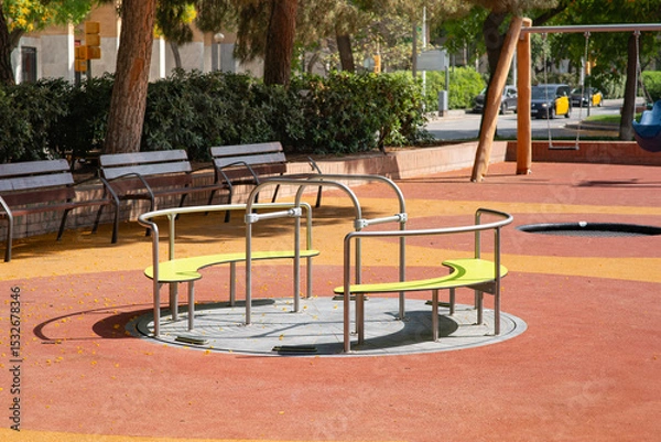 Obraz Circular playground carousel with lime green seats and metal bars on a cushioned, red rubber surface. Nearby, several wooden benches are positioned along the perimeter. Nobody