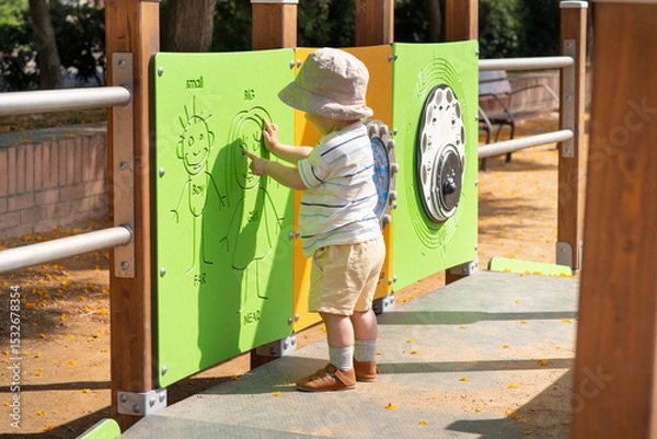 Obraz A baby boy wearing a sun hat, striped shirt, and shorts explores a playground. He is interacting with a green activity panel featuring simple drawings designed for sensory play. 