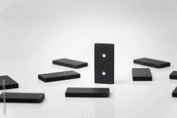 Fototapeta A black domino piece stands upright amidst others lying flat on a reflective white surface.