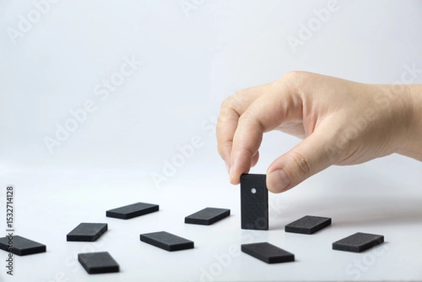 Fototapeta Hand arranging a domino piece within scattered tiles on a plain white surface, portraying concepts of strategy, decision-making, and sequencing processes.