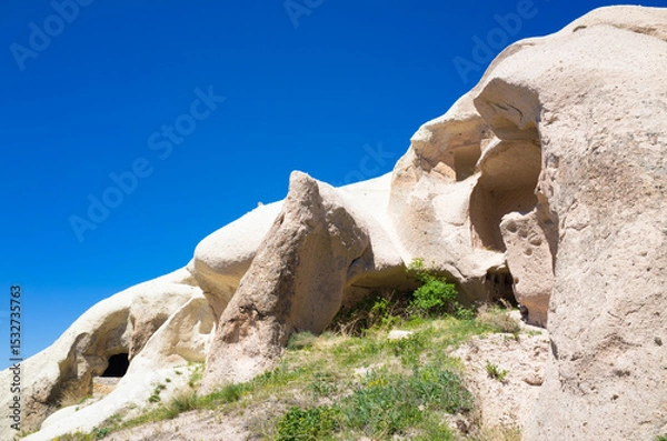 Obraz Natural rock formations with ancient cave dwellings carved into soft tuff rock in Cappadocia, Turkey. Scenic summer landscape of Cappadocia with arid terrain, bright blue sky and sparse vegetation.