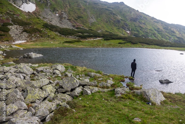 Fototapeta Outdoor enthusiast trekking across stone-covered lakeside in remote wilderness. Dramatic mountain scenery with cloudy sky and pristine water.