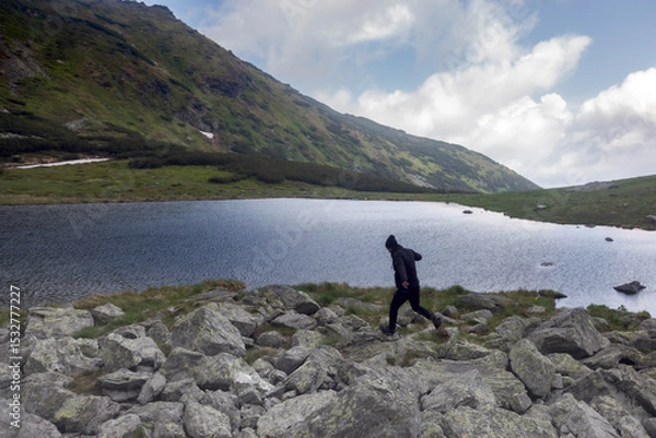 Fototapeta Hiker celebrating freedom by jumping across alpine brook, golden flowers framing scenic glacial valley with steep rocky slopes and snowfields