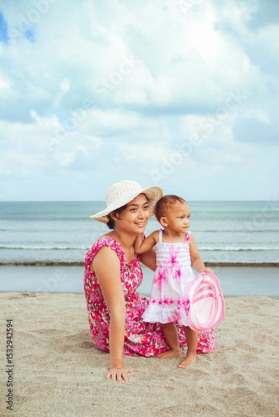 Fototapeta Asian mom and toddler girl in beach dresses having fun together during vacation at a tropical beach on a summer day with blue ocean and cloudy sky on the background